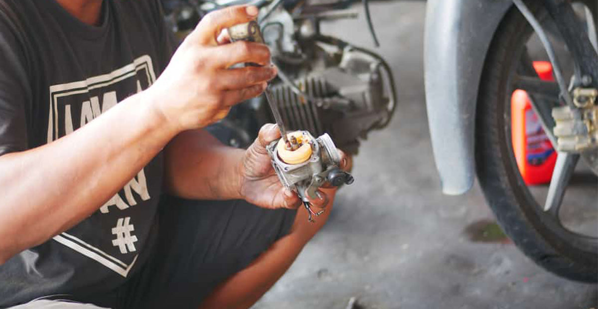 The Man is Squatting and Using a Screwdriver to Take Apart a Motorcycle Carburetor